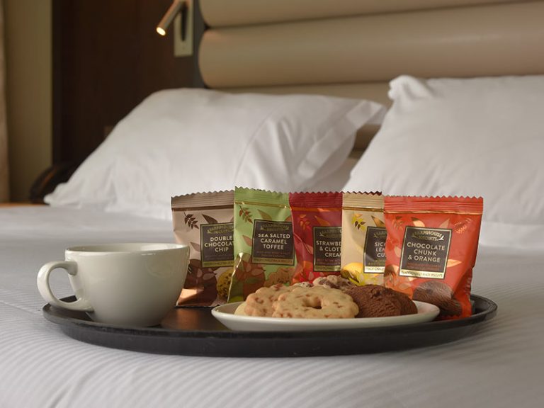 Hospitality tray placed on hotel bed. Tray has a cup of Tea or coffee and a plate with hotel Biscuit packets laid out for guests. Individually wrapped biscuits sit behind the plate ready for replenishment.